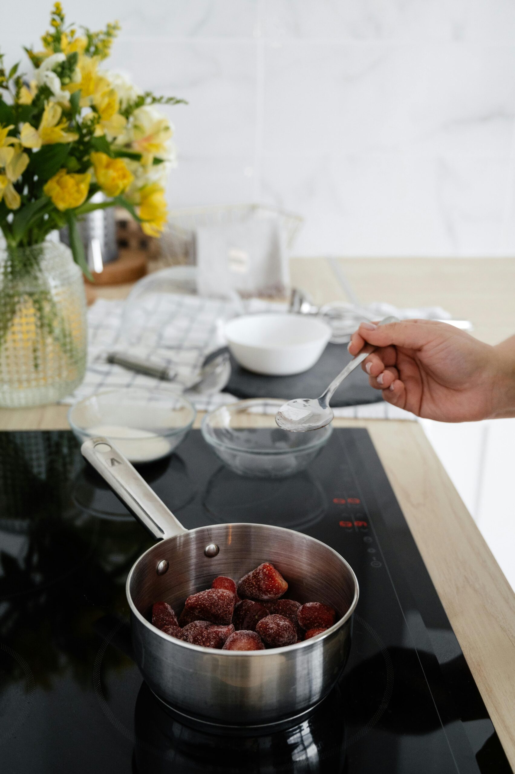 Female hand putting starch into bowl with frozen strawberry on stove while preparing confiture in bright kitchen